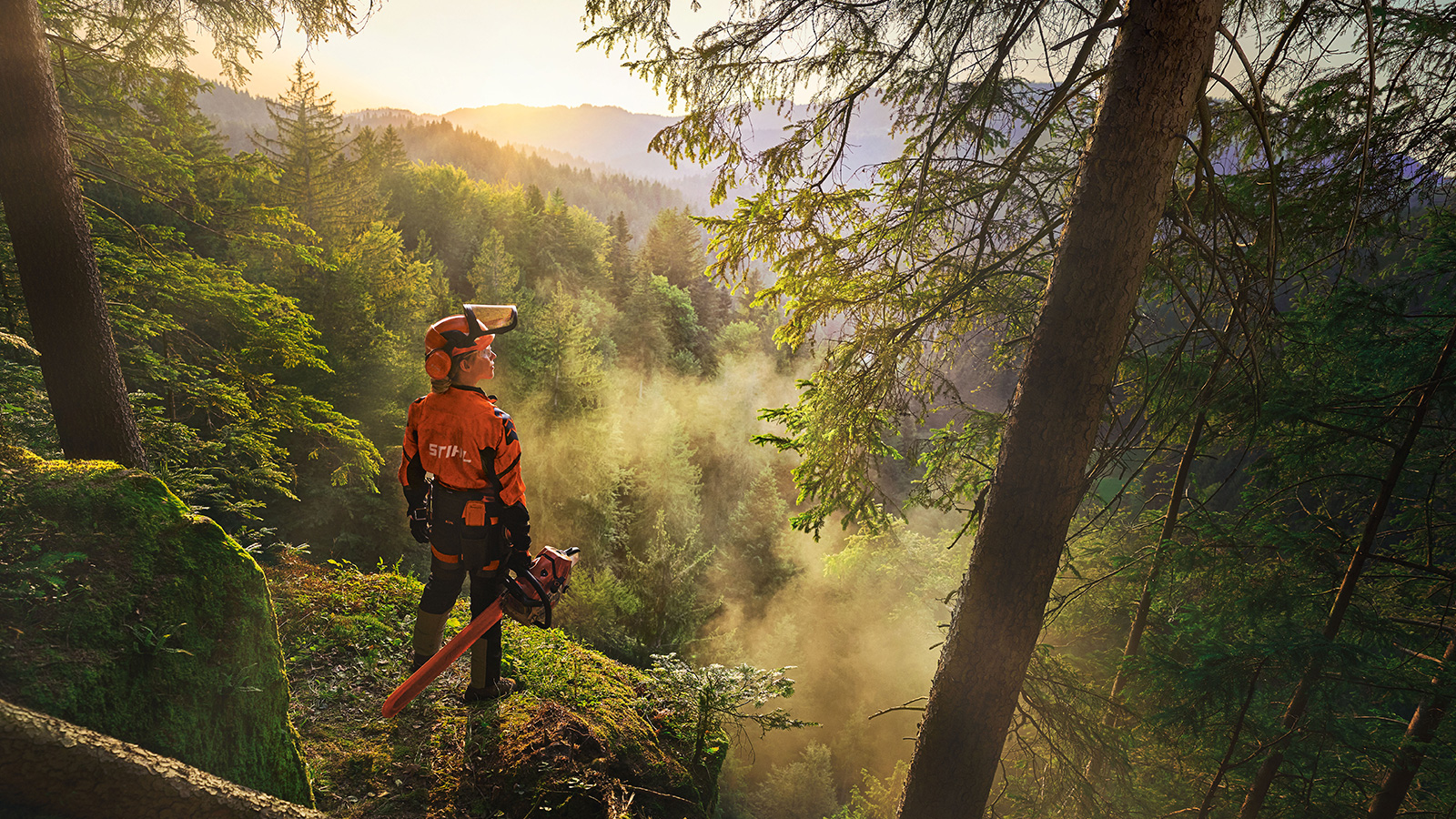 A woman is standing at the edge of a cliff in a saturated green forest. She is wearing orange STIHL workwear, the ADVANCE ProComHeadset, and holding a STIHL MS Chainsaw.