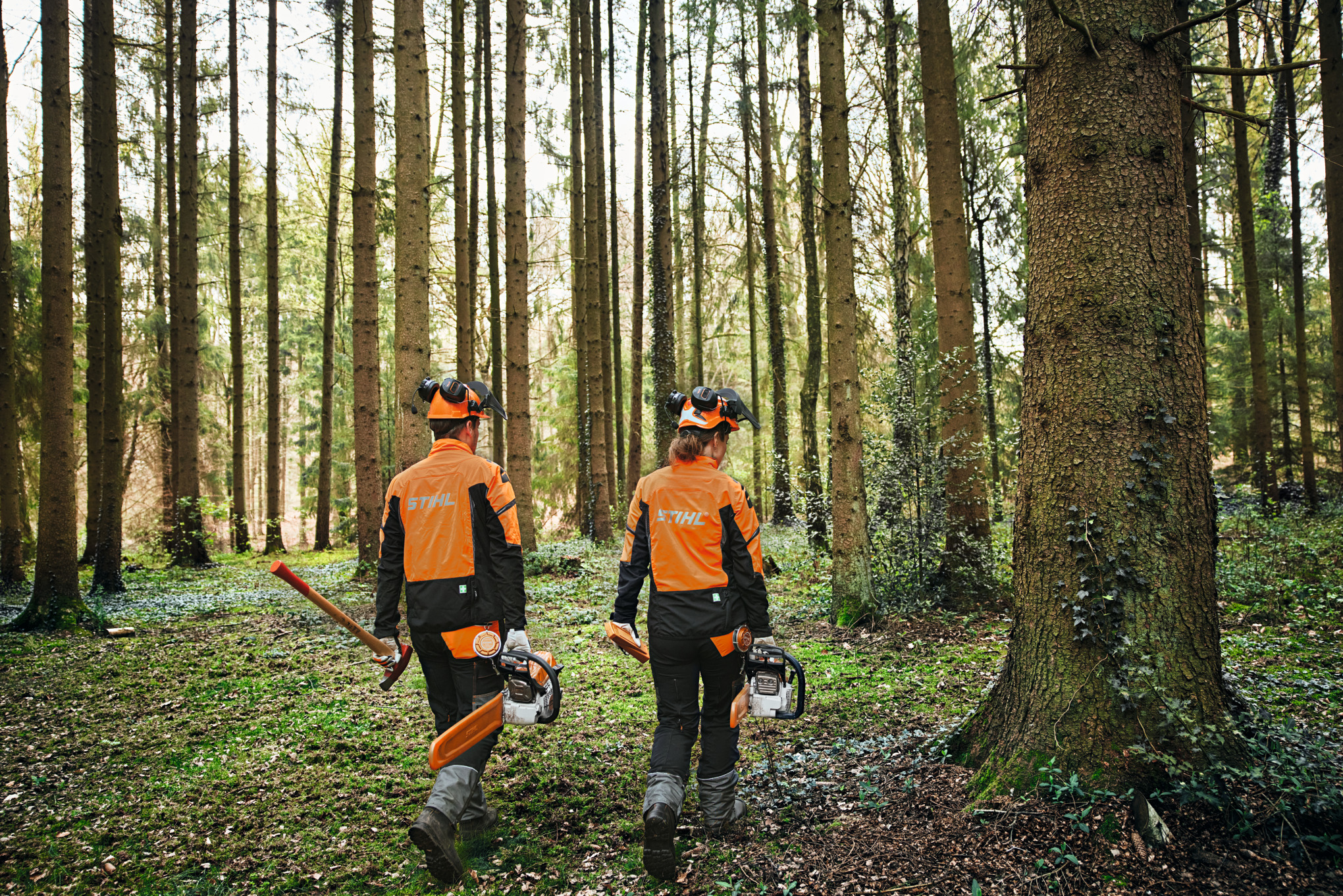 Two people wearing protective equipment in a forest, with chainsaws and axe.