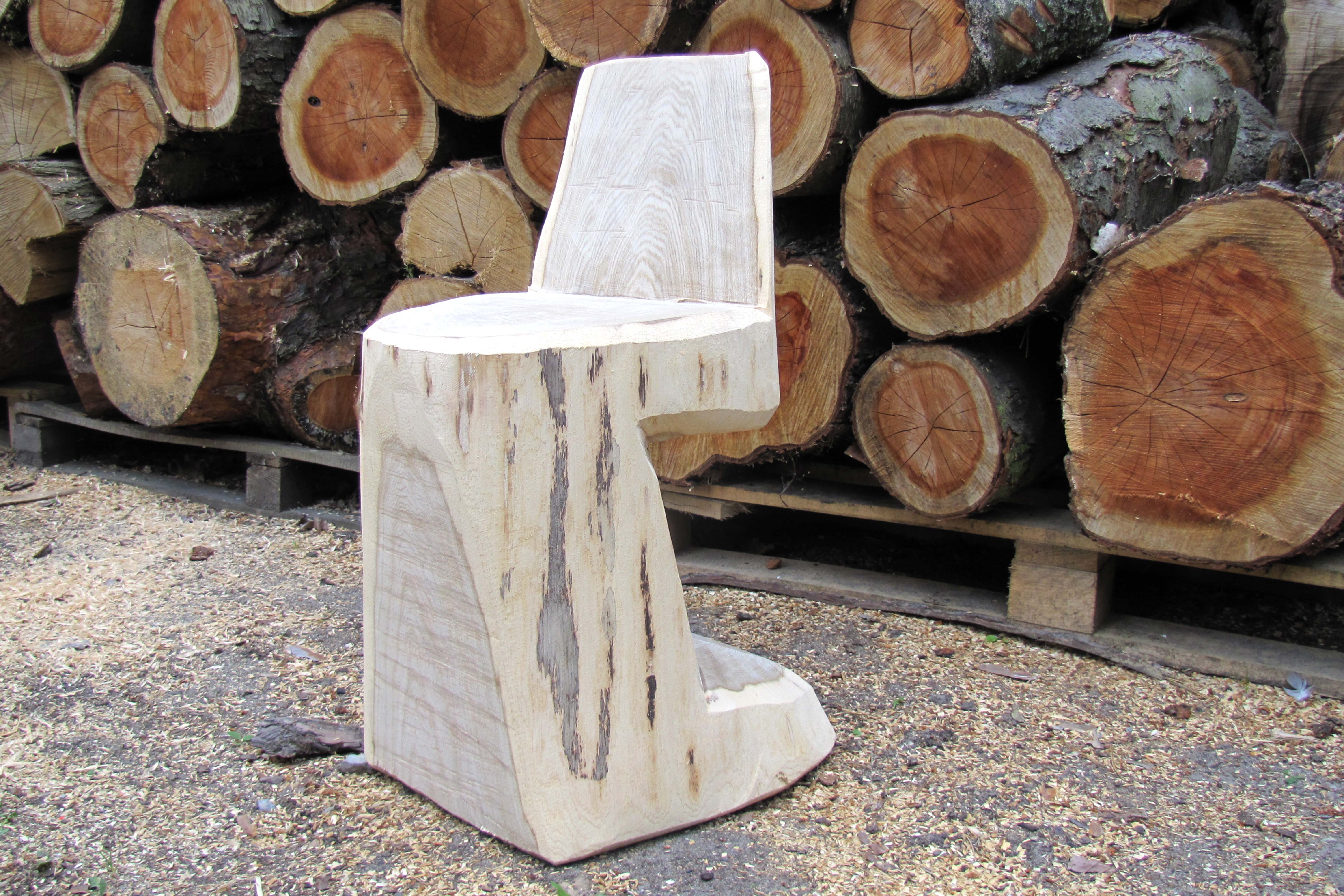 A finished log chair in front of a pile of tree trunks