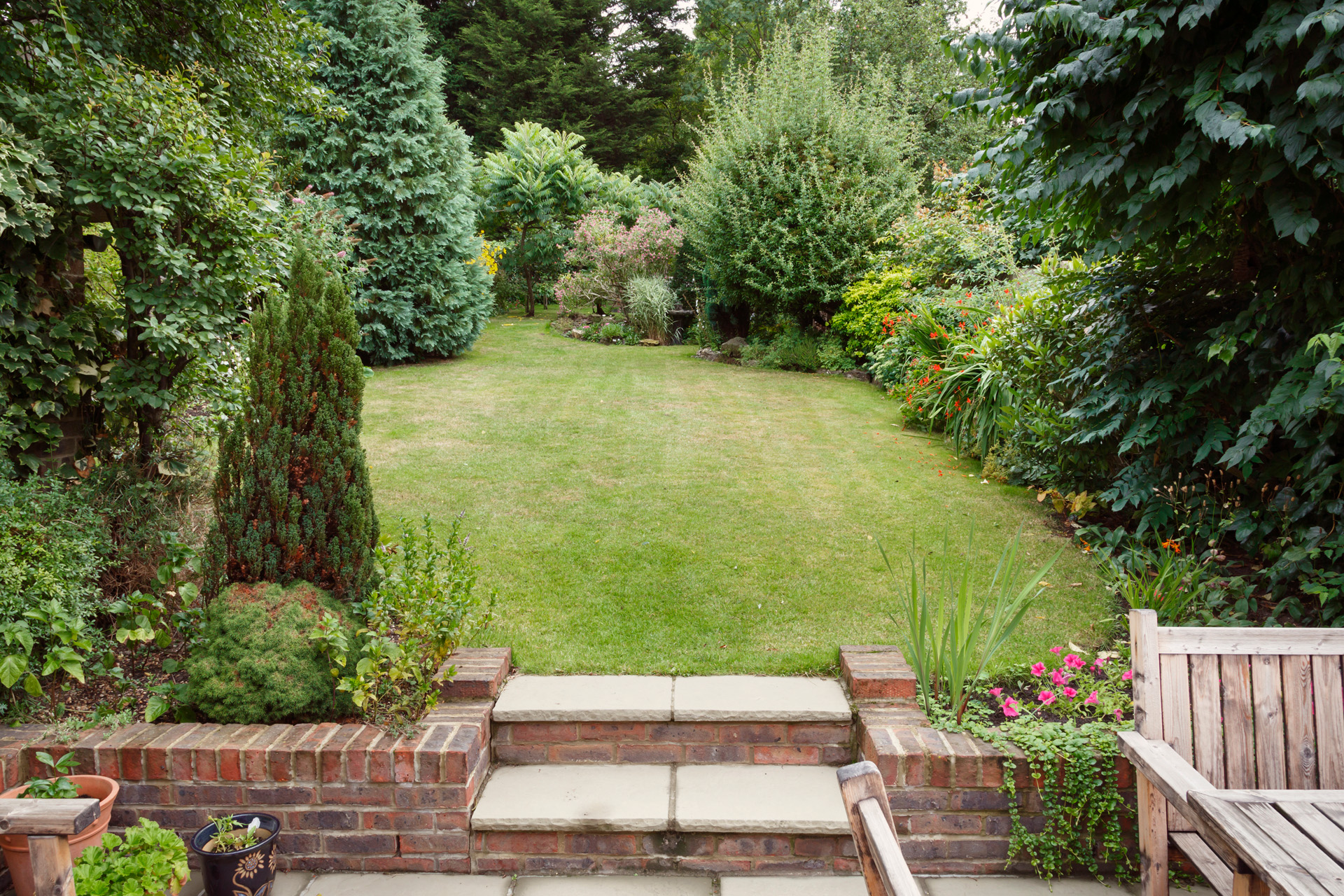 Small garden with patio furniture and plant tubs in the foreground and a lawn with small garden trees and shrubs behind.