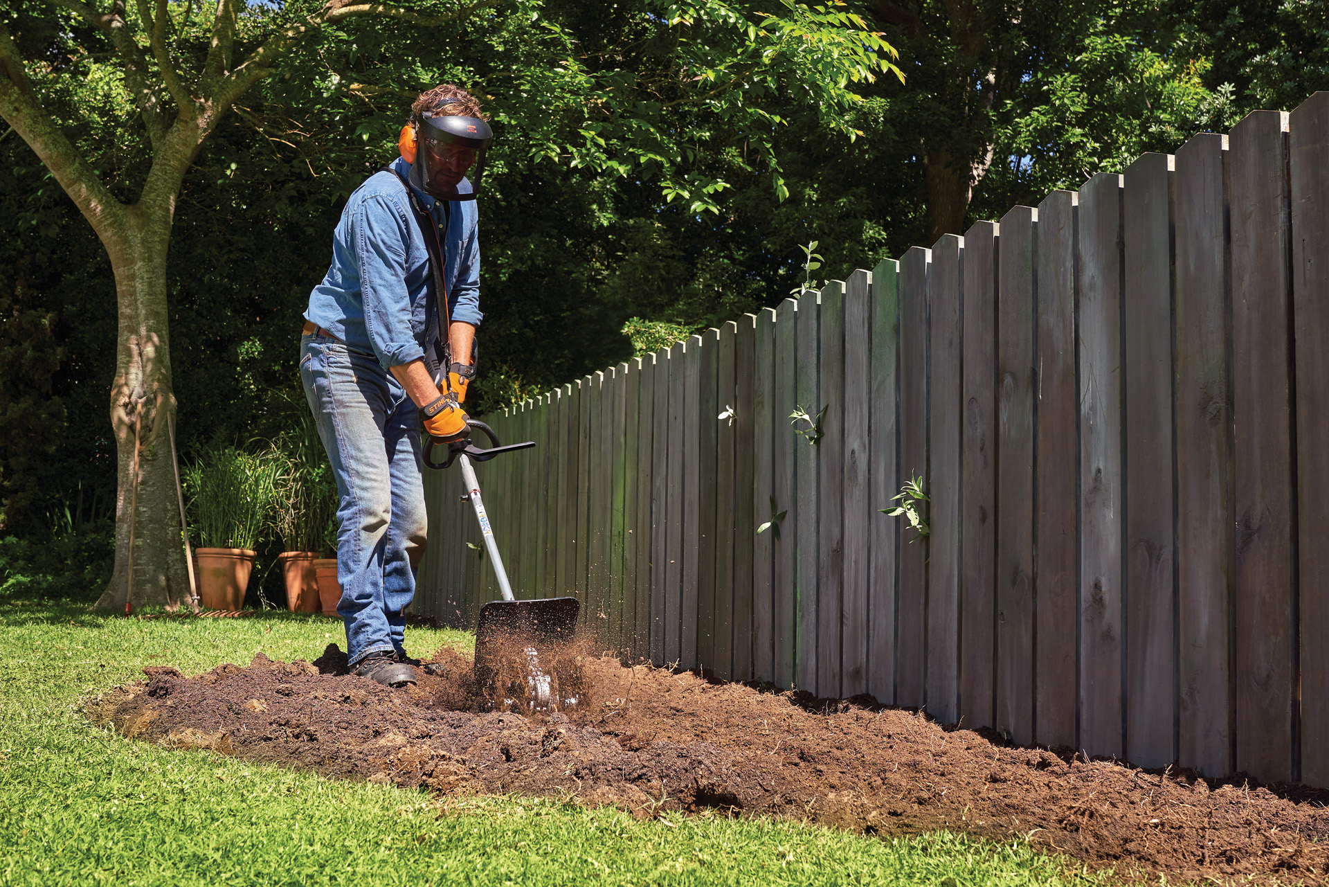 A person wearing personal protective equipment cultivates soil in a garden using a STIHL KombiSystem with pick tine
