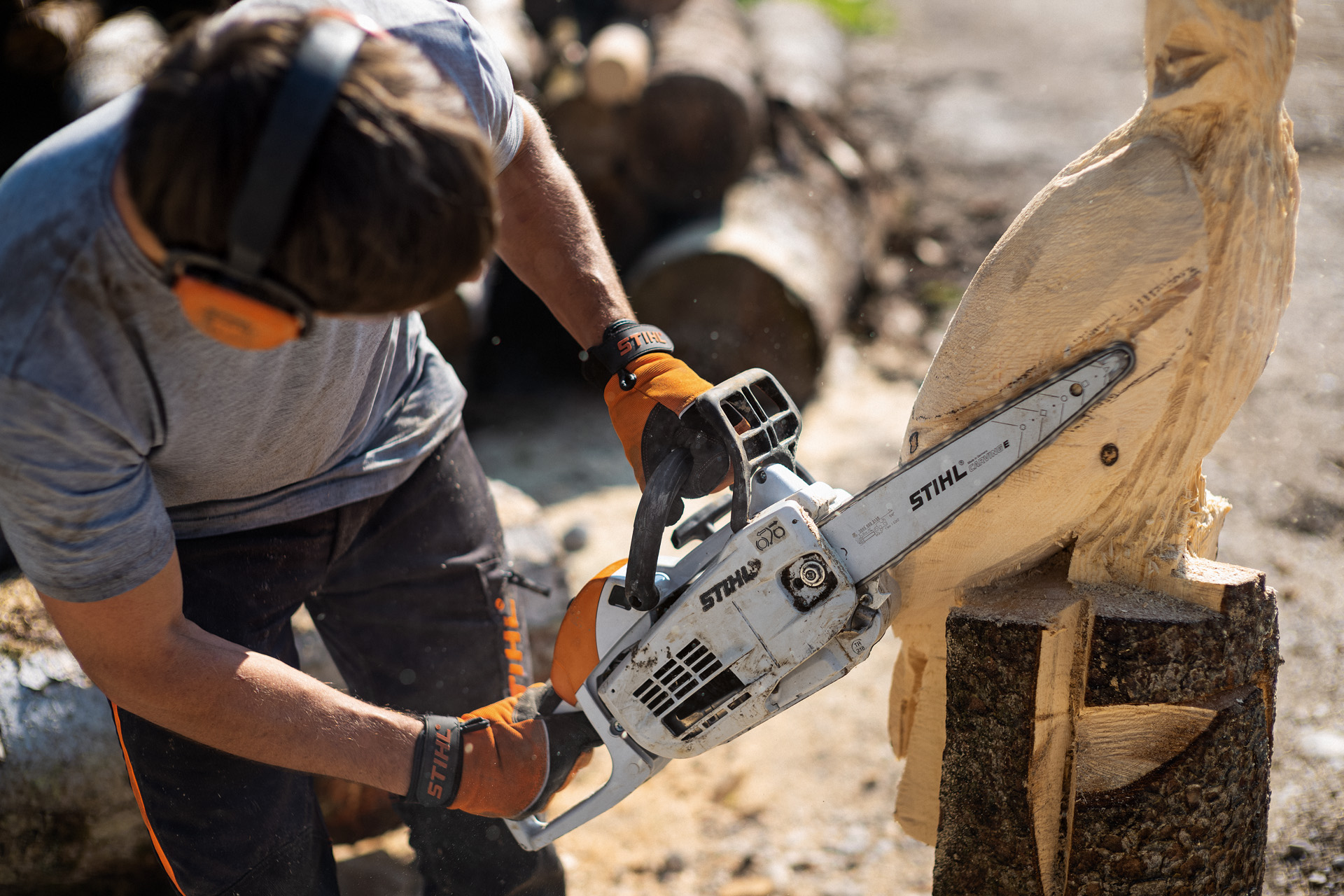 A man wearing ear protection and gloves uses a STIHL MS 193 C-E carving chainsaw to work on a partly finished wooden sculpture of a bird