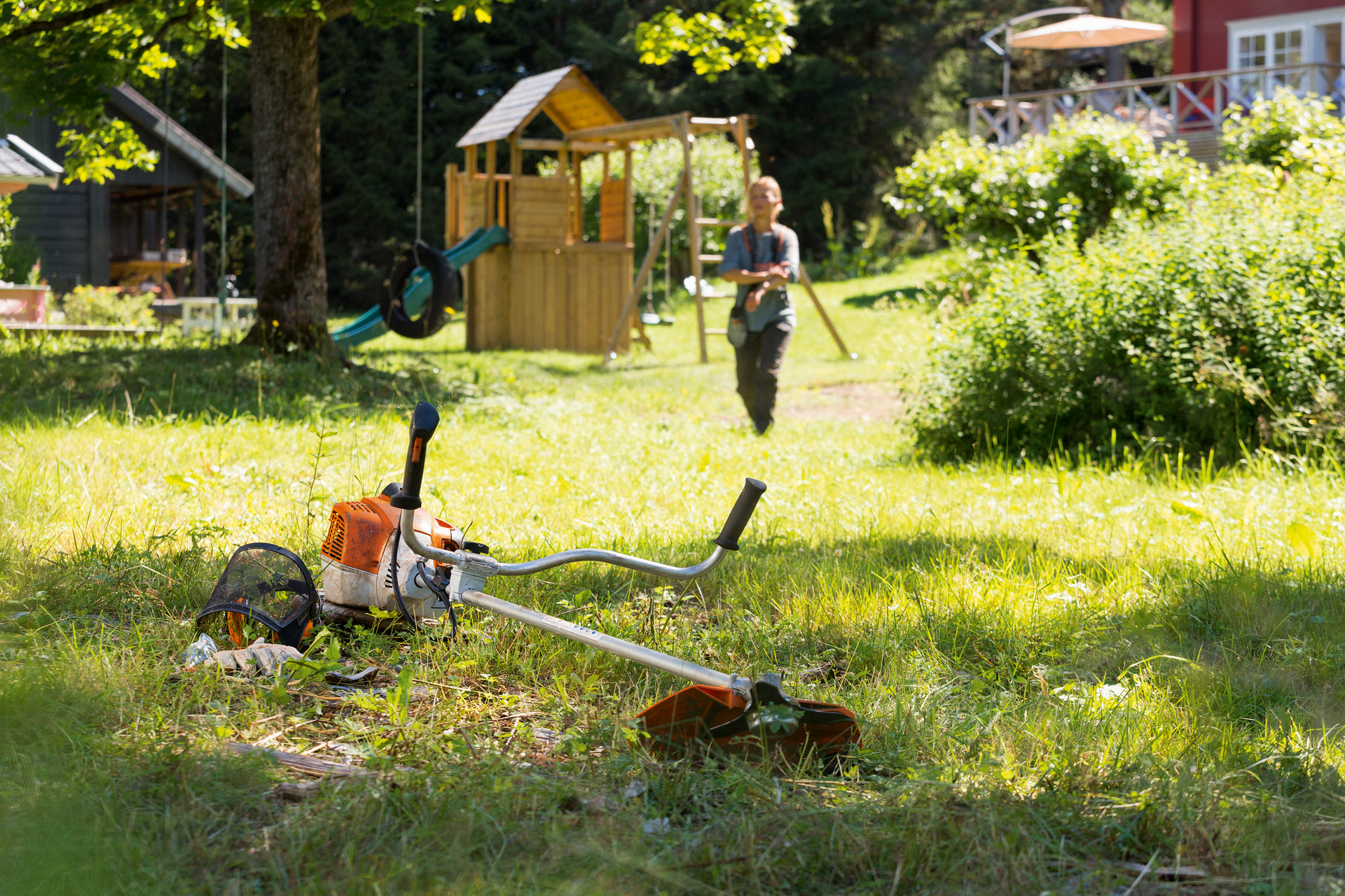 A STIHL FS 240 brushcutter lies on the ground in a yard, with play equipment and a person approaching in the background