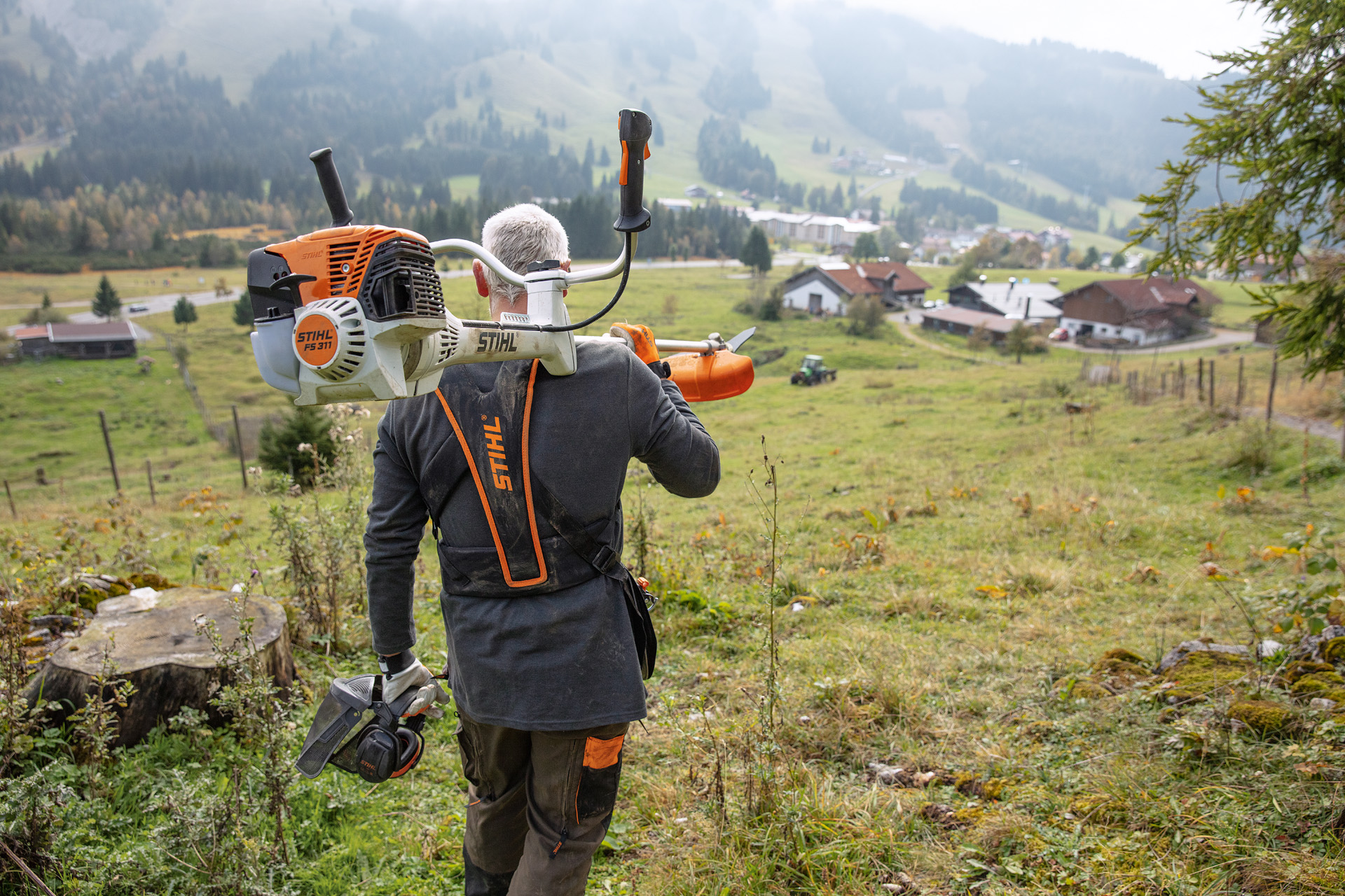 A man is walking away with a STIHL FS311 clearing saw on his shoulder and in the background is a green hill and houses 