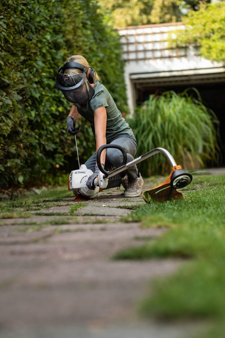 Woman wearing PPE starting a STIHL FS 38 petrol grass trimmer on garden path