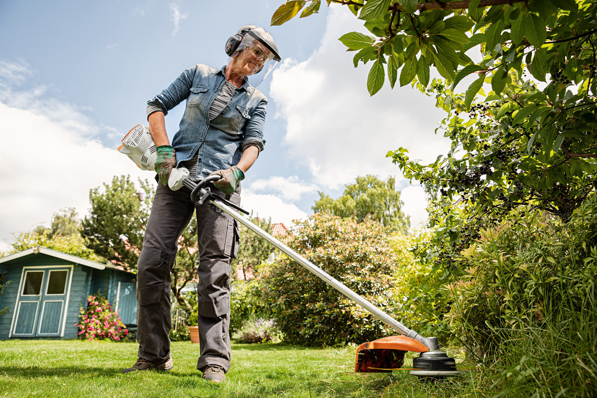 A woman wearing face and ear protection using a STIHL FS 56 R petrol grass trimmer in a garden with shrubs and lawn