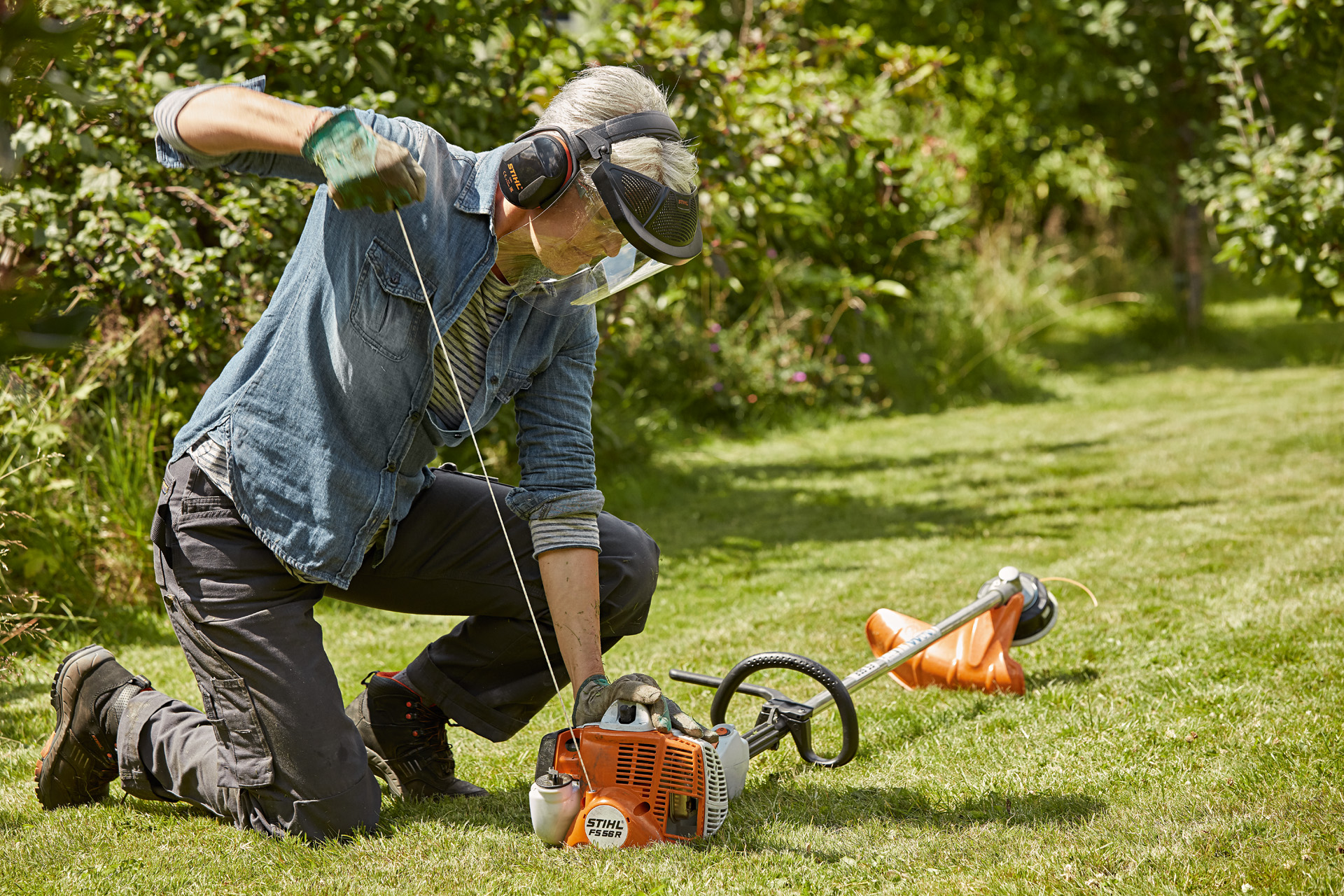 A person wearing ear and face protection starting a STIHL FS 56 R petrol grass trimmer on a garden lawn