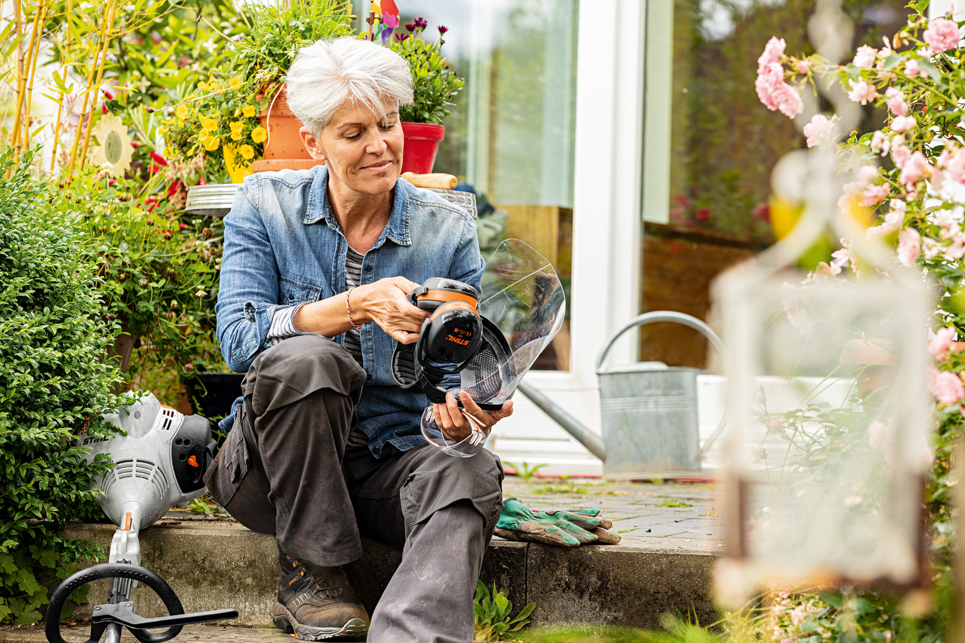 A woman sitting on a garden step, holding a STIHL face visor with ear defenders, with a grass trimmer laying nearby