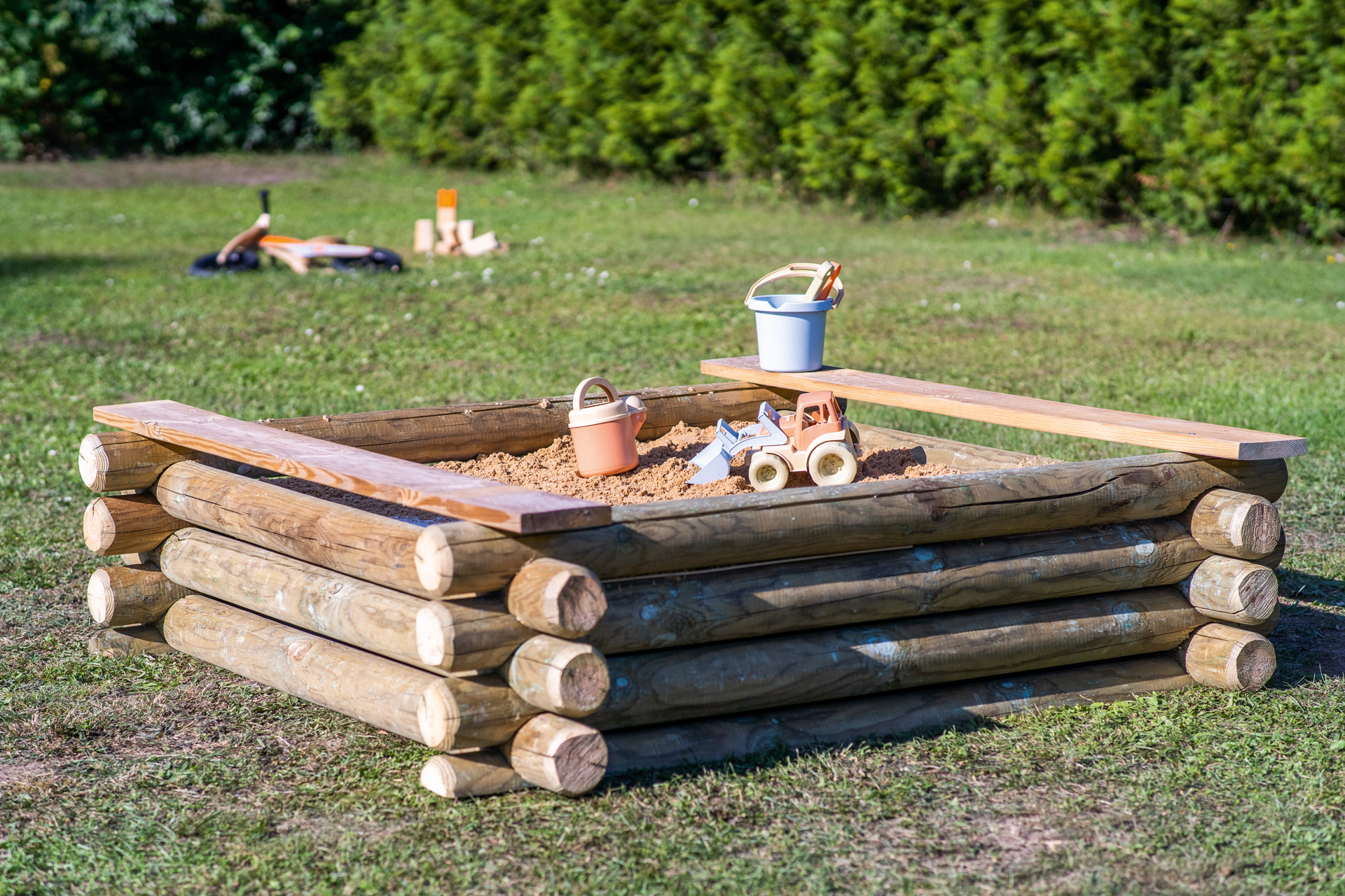 A wooden sandpit with bench seats stands on a green lawn, with buckets and other toys inside it.