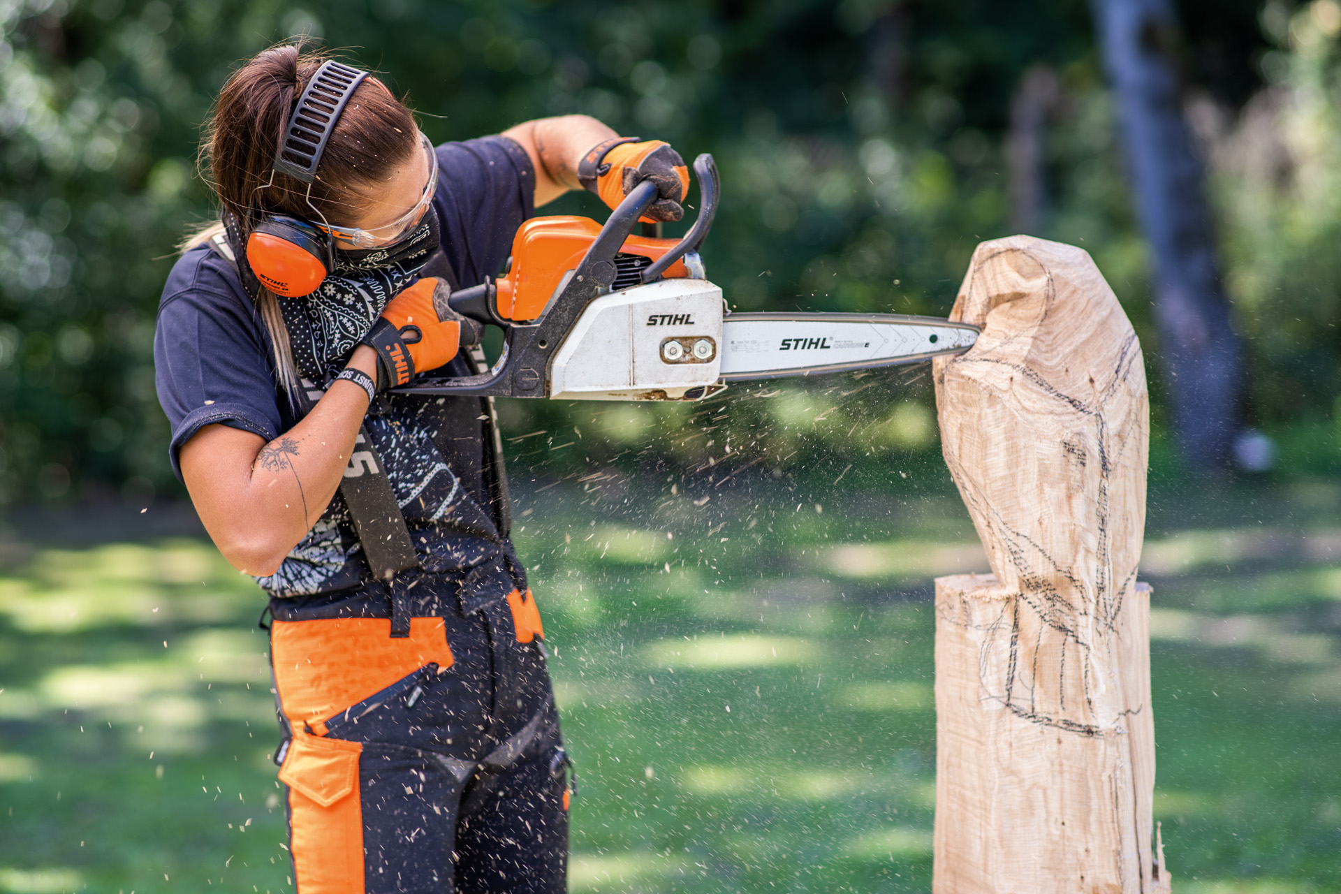 A woman wearing protective equipment carving a wooden owl with a chainsaw