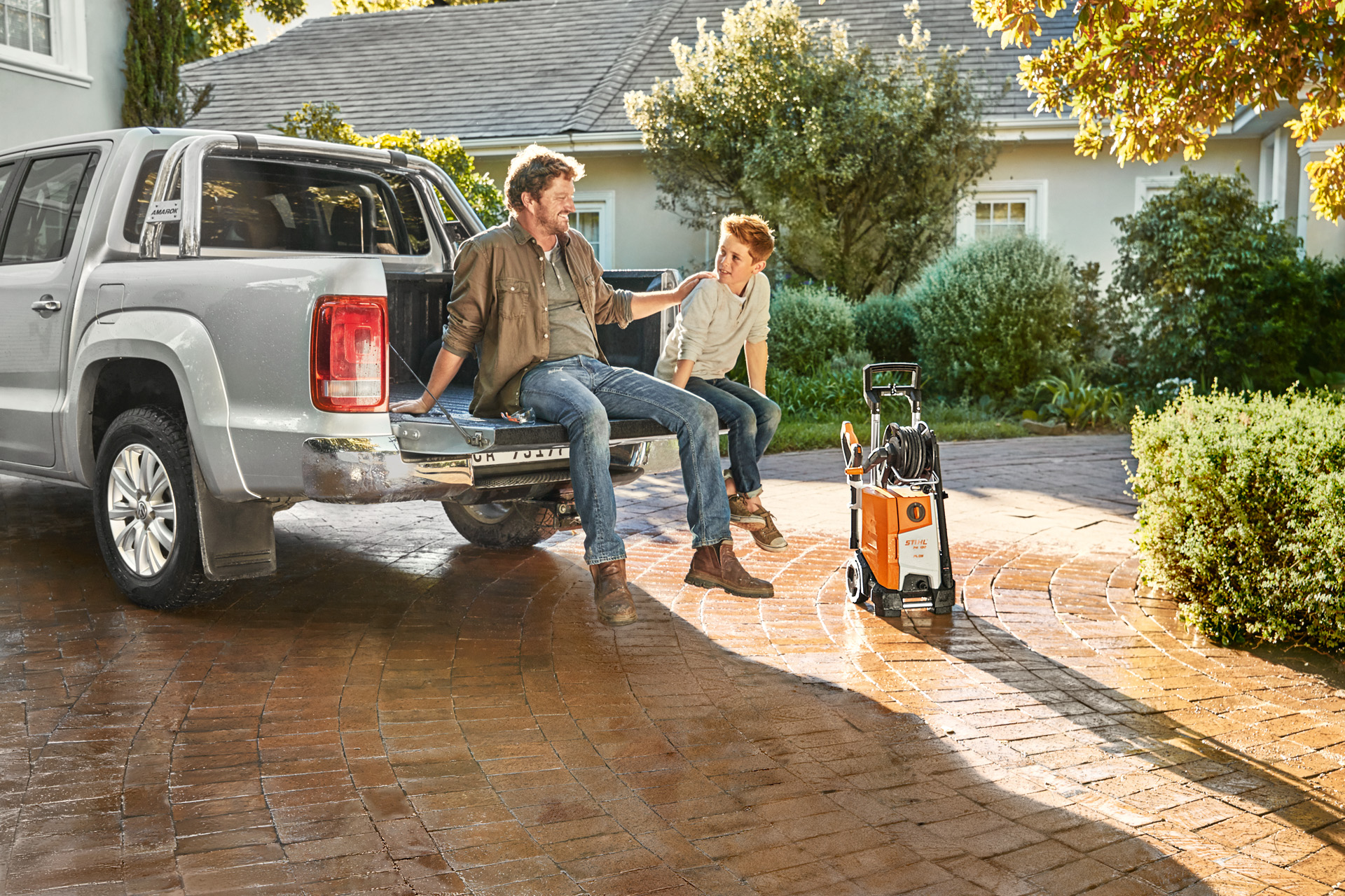 A man and young boy sit on the truck bed of a pick-up, with a STIHL RE 130 plus high-pressure cleaner beside the vehicle