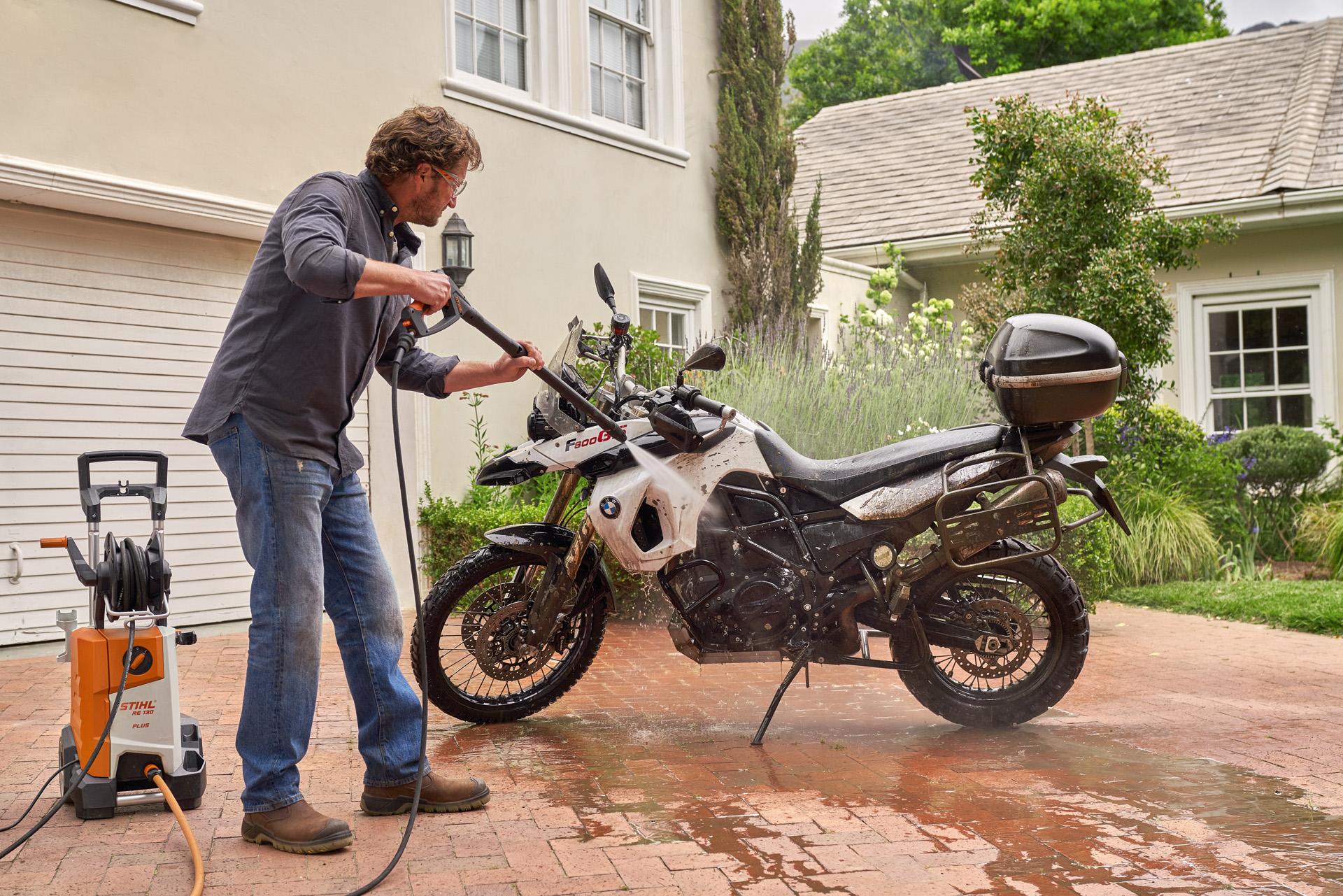 A man using a STIHL RE 130 Plus high-pressure cleaner to wash a white motorcycle on a home driveway.