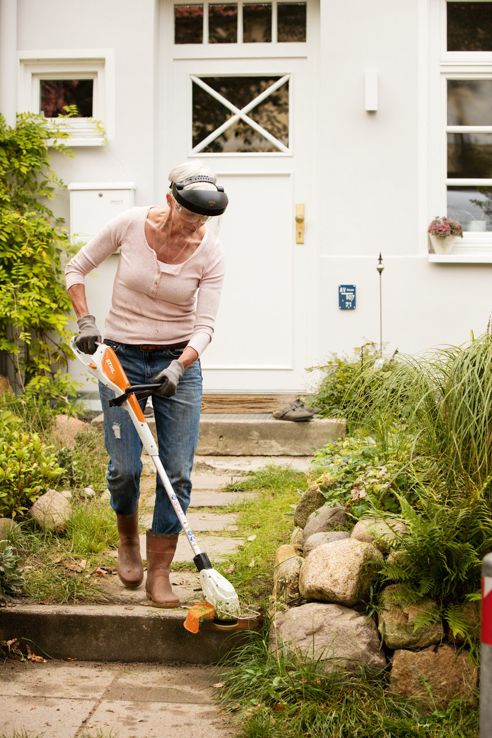 Woman wearing safety glasses and work gloves trims lawn edging along stone path with STIHL FSA 45 cordless lawn trimmer.