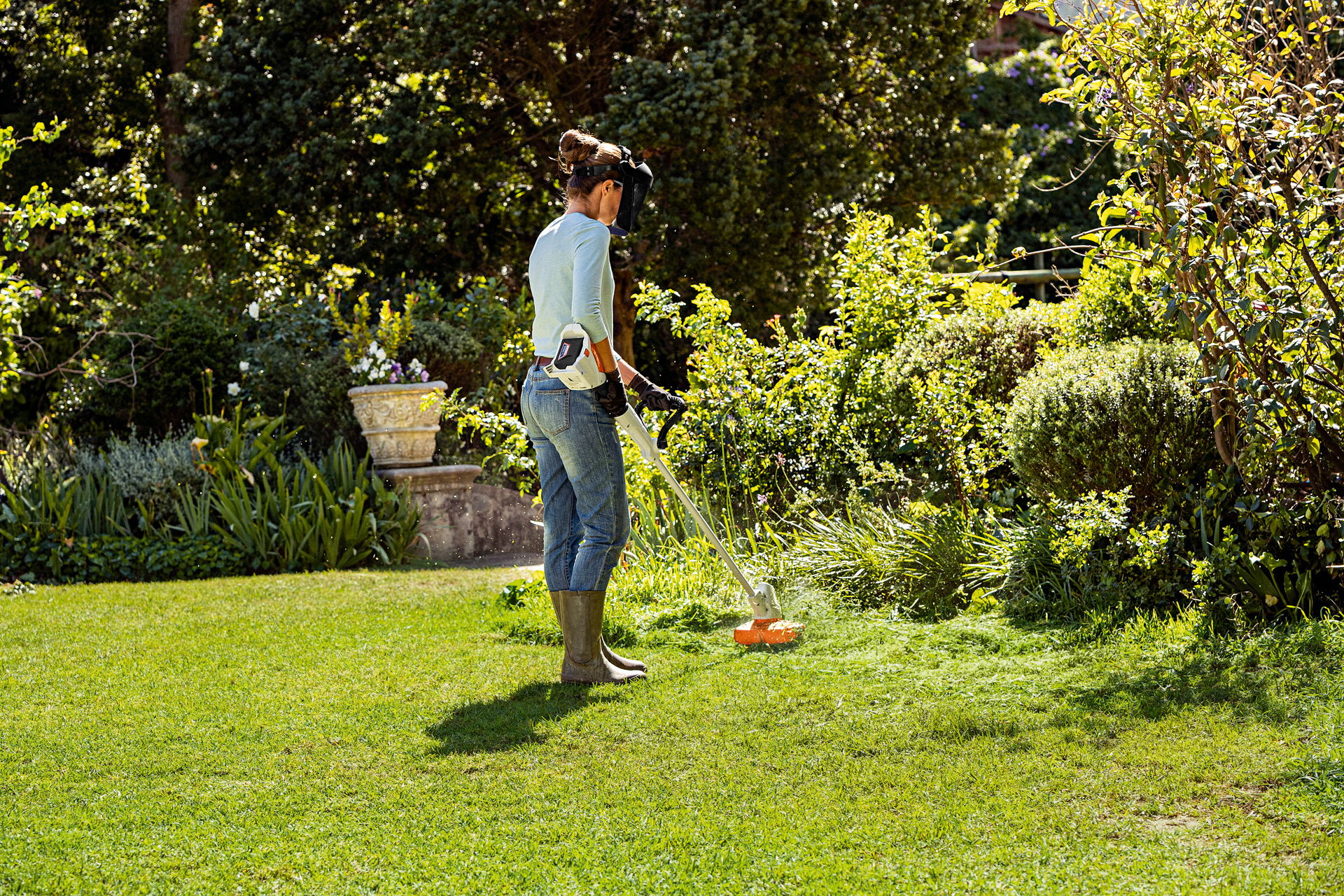 A woman wearing a face guard and gloves using a STIHL FSA 56 cordless grass trimmer in a garden