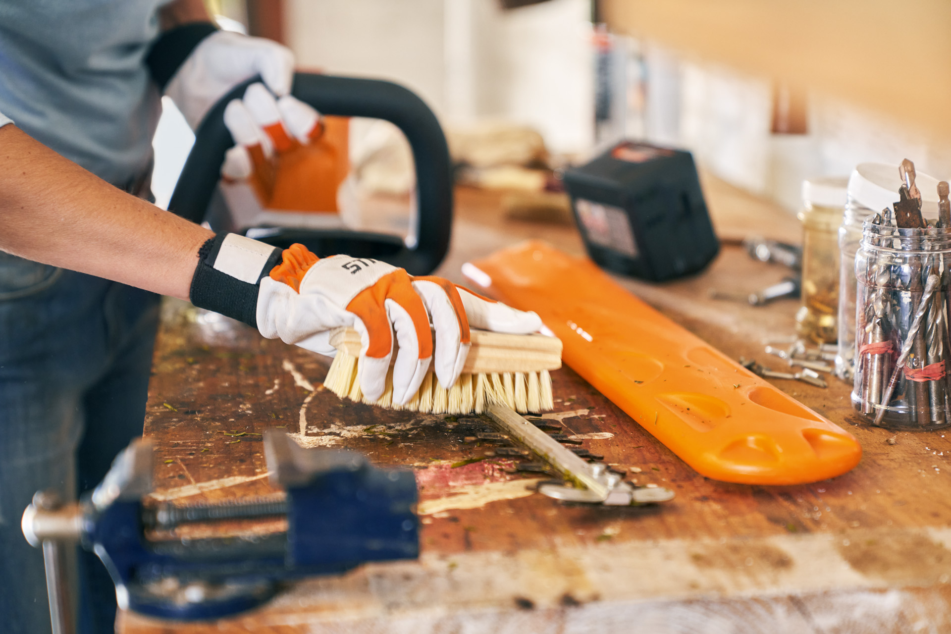 A STIHL HSA 56 cordless hedge trimmer on a bench, with gloved hands cleaning the cutting attachment with a brush