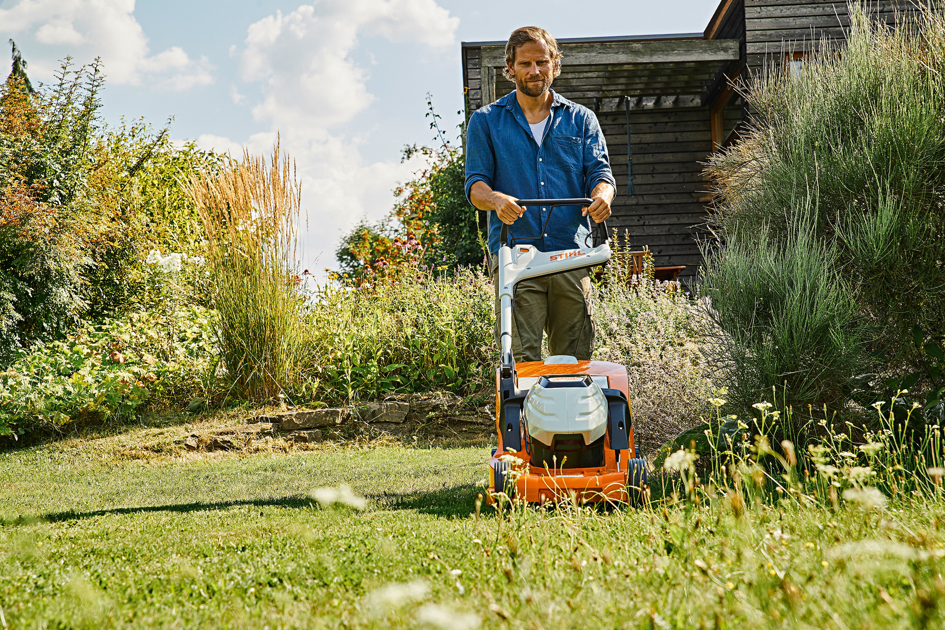 A man is halfway through mowing untidy and overgrown grass using a STIHL RMA 448 P-C cordless lawn mower, in a garden surrounded by tall shrubs and plants