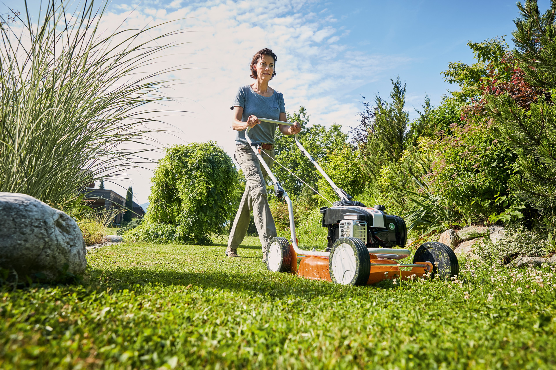 A woman using a STIHL RM 2 R petrol mulching mower on a lawn bordered with bushes and grasses