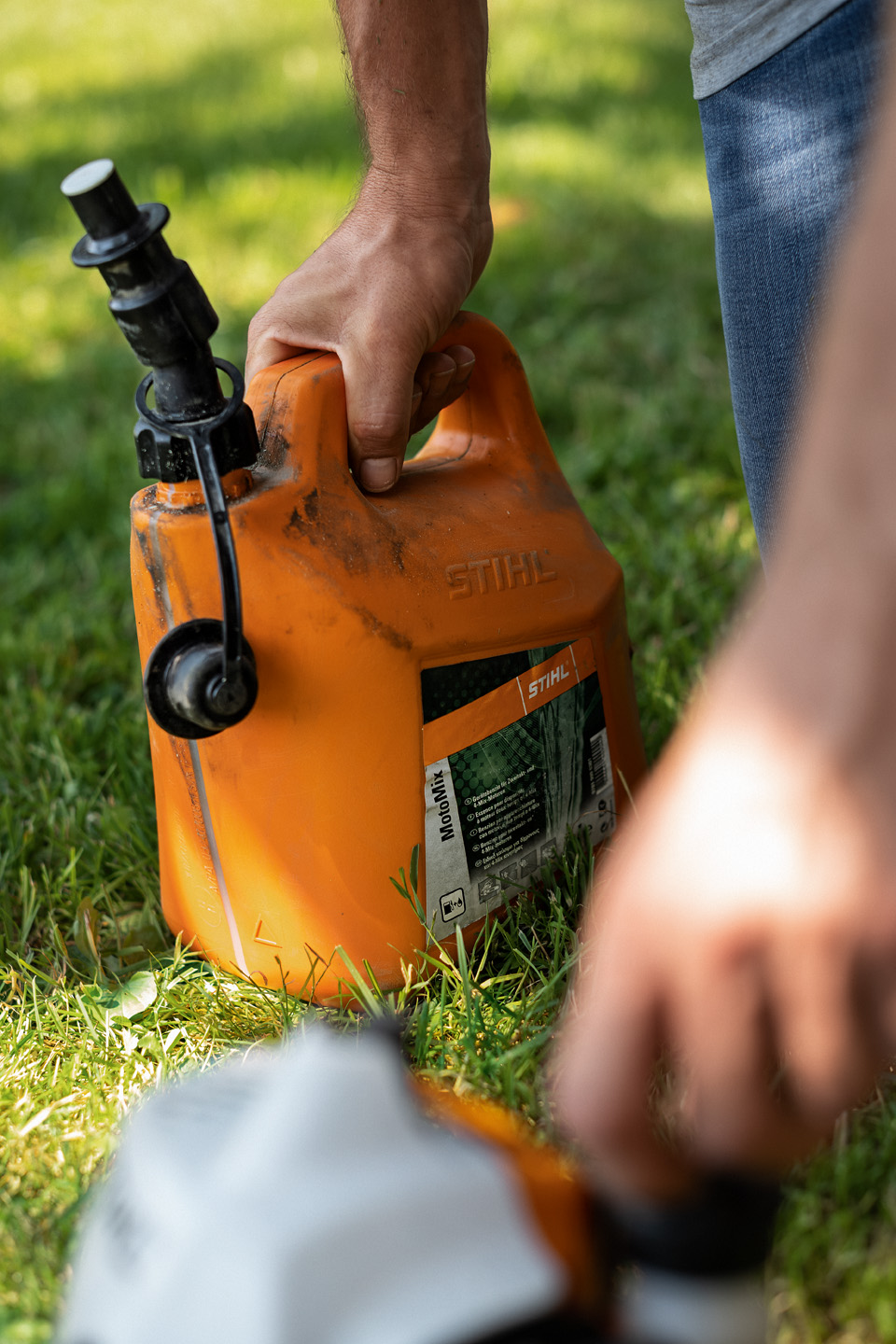 A dirty canister of STIHL MotoMix fuel mix on some grass, with a hand opening the fuel cap of a tool in the foreground