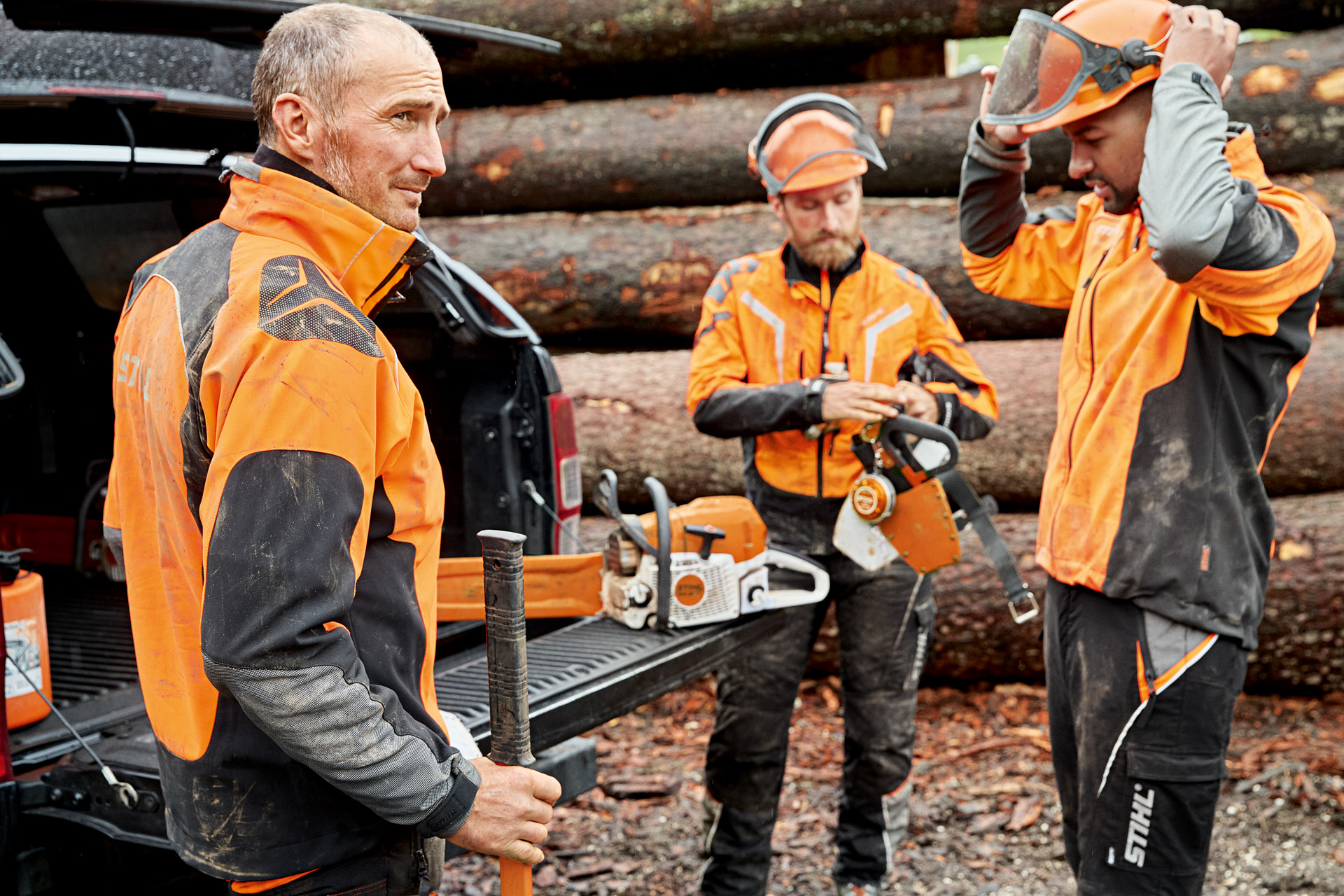 Three men wearing STIHL protective clothing prepare to work with a STIHL chainsaw with a stack of tree trunks behind