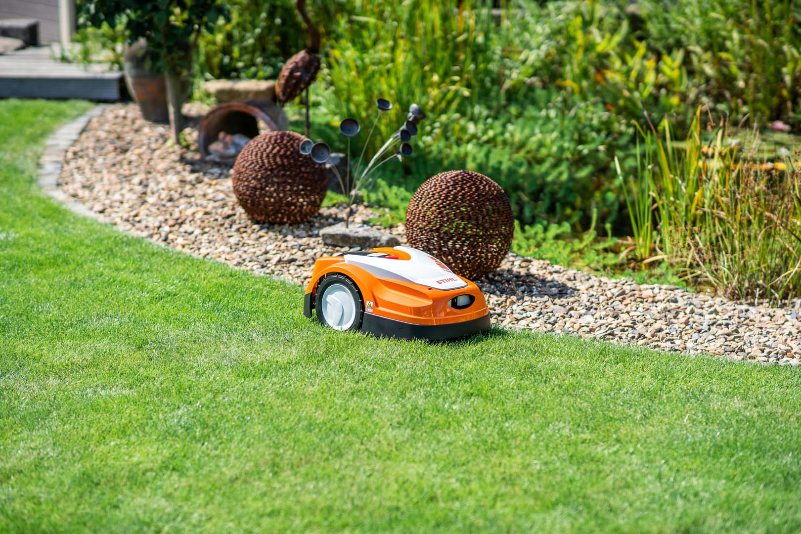 A STIHL iMOW® 422 robot mower on a lawn beside a gravel bed with garden sculptures