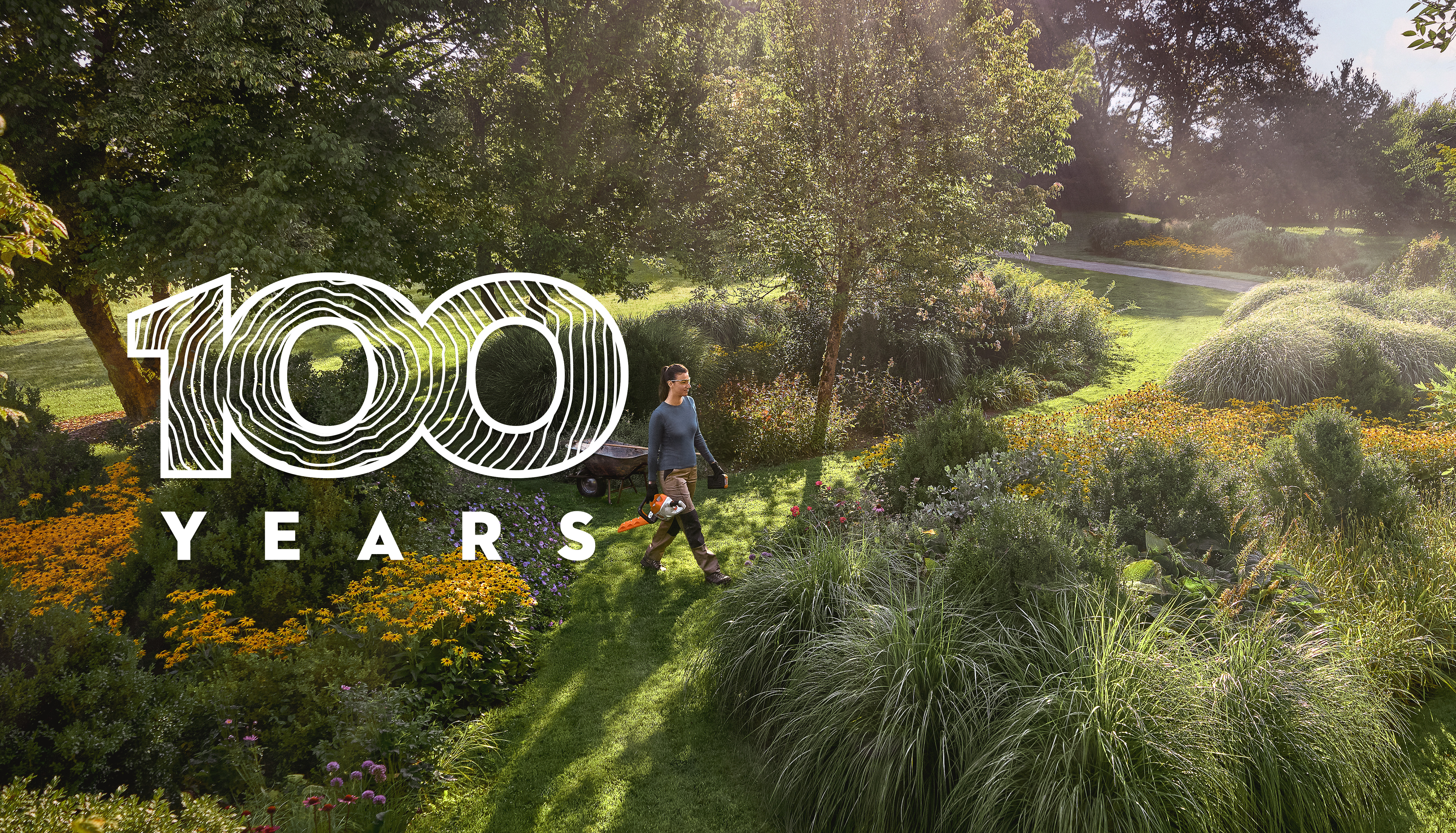 100 years graphic in front of woman working with hedge trimmer in flowering landscape garden on sunny day